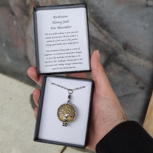 Hand holding an open black gift box with a Tree of Life pendant over a golden honey jade disc on a chain, and a card reading Birthstone Honey Jade For November