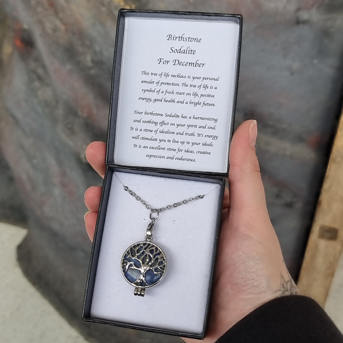 Hand holding an open black gift box with a sodalite Tree of Life pendant on a chain and a card reading Birthstone Sodalite For December