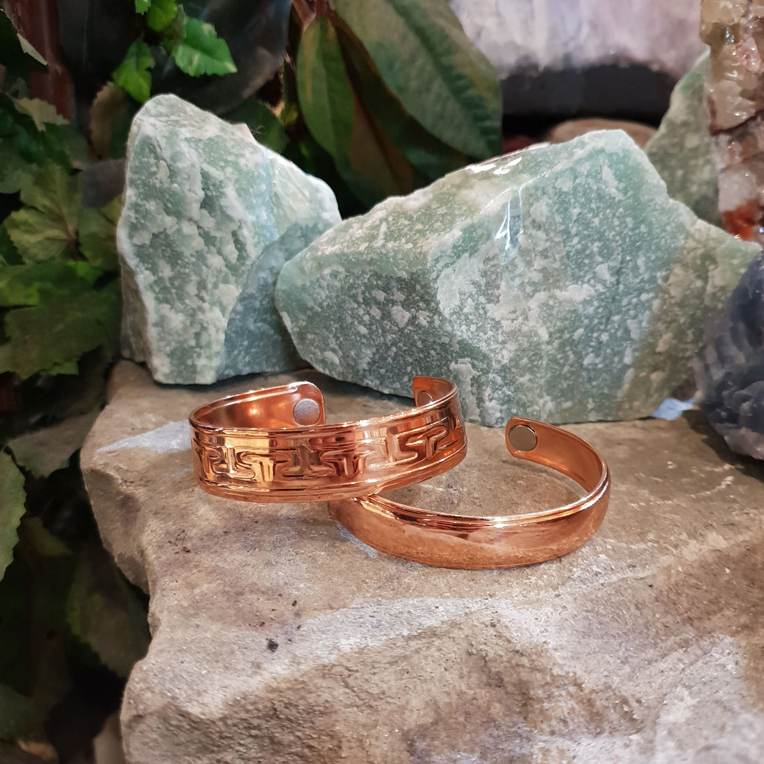 Two copper cuff bracelets, one patterned and one plain, resting on a stone slab in front of rough green aventurine