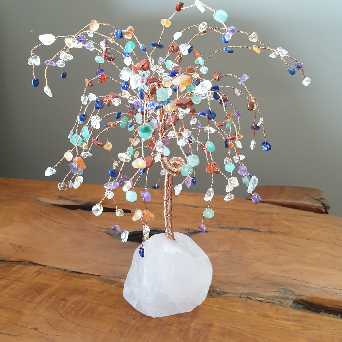 Wire crystal tree with small multicoloured gemstone leaves on a white rose quartz base, standing on a wooden table