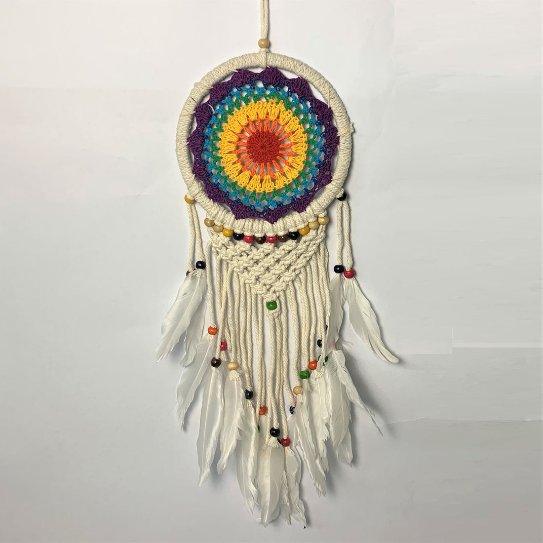 White crochet dreamcatcher with a rainbow centre, beads and white feathers hanging against a pale wall