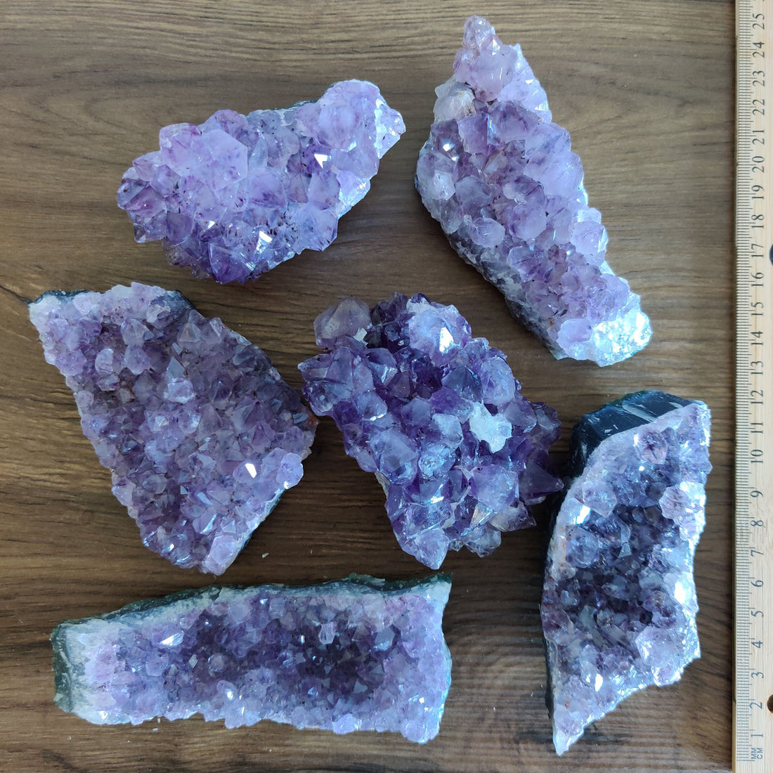 Six natural amethyst crystal clusters laid on a wooden table beside a ruler, their purple points catching the light