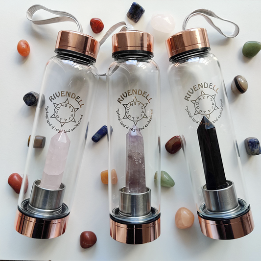 Three glass crystal water bottles with rose-gold lids holding rose quartz, amethyst and black obsidian points, laid among tumbled crystals