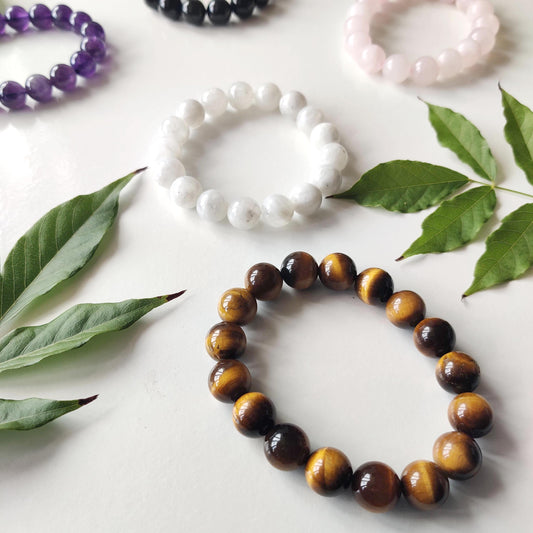 Round-bead crystal bracelets in tiger's eye, amethyst, black stone, rose quartz and white stone laid on a white surface with green leaves
