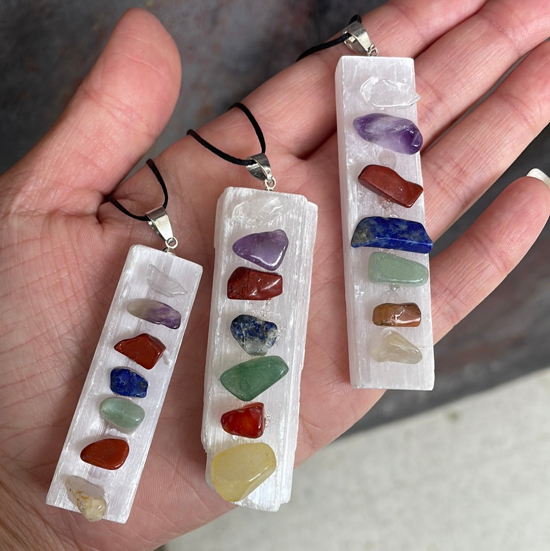 Three white selenite bar pendants on black cords, each set with seven small tumbled chakra stones, held in an open hand