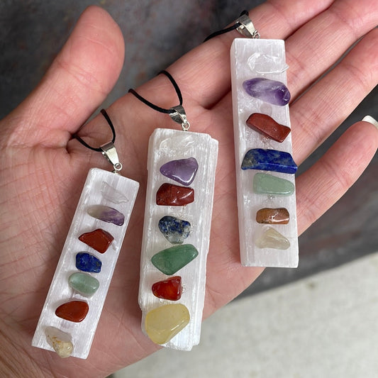 Three white selenite bar pendants on black cords, each set with seven small tumbled chakra stones, held in an open hand