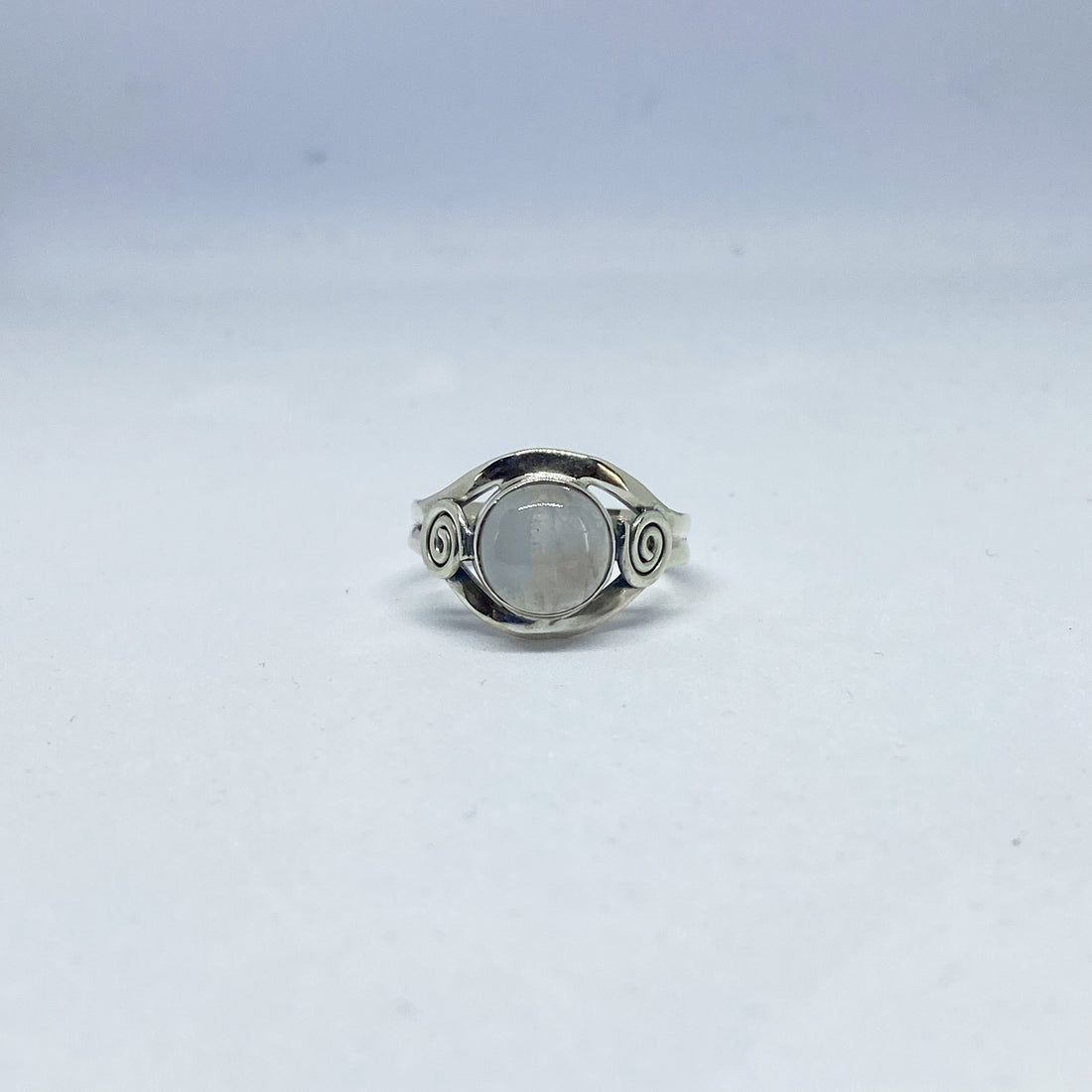 A round moonstone set in a 925 sterling silver ring with a silver spiral on each side, on a pale surface
