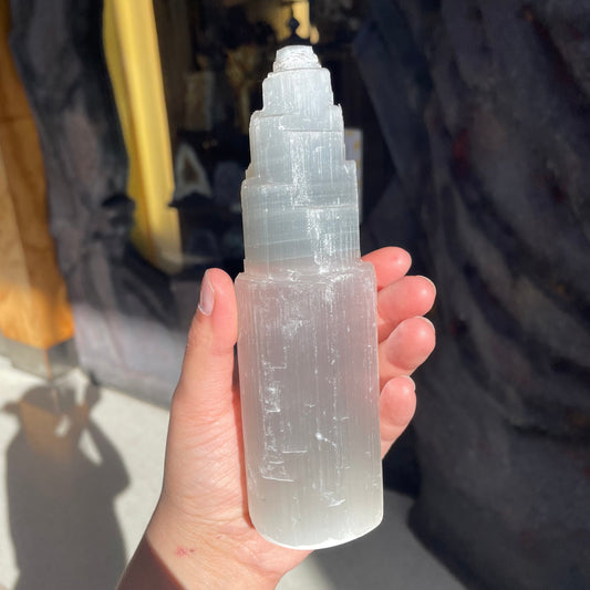 Hand holding a white selenite tower with stepped top in warm sunlight inside the shop