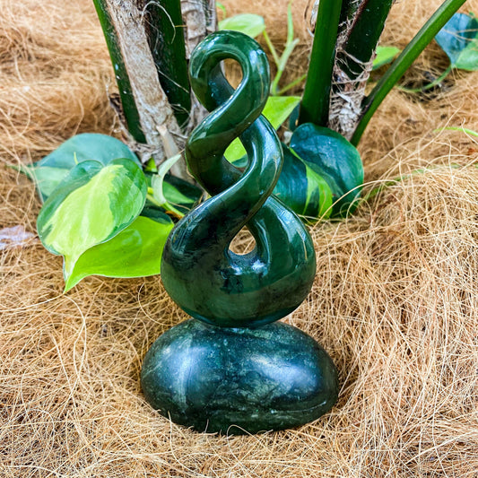 Deep green greenstone double twist carving standing on a rounded greenstone base among green leaves and coconut fibre