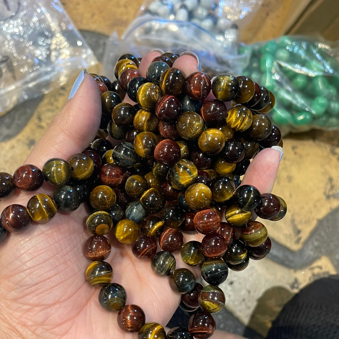 A hand holding strands of polished tiger's eye bead bracelets in gold, brown, red and blue tones