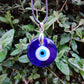 Blue glass evil eye pendant on its black cord against green leaves